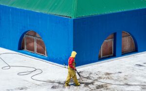 A worker in protective gear power washes a vibrant blue building exterior.
